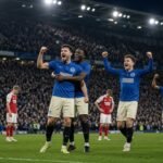 Create a professional, realistic football news featured image for an article titled “Why Brighton Beat Arsenal 3-0”. Show a dramatic Premier League match atmosphere at the Amex Stadium. Feature Brighton players celebrating passionately after scoring, with Arsenal players looking disappointed in the background. Include the Brighton and Arsenal team colours naturally through the players’ kits, but do not use official logos unless necessary. Show a packed stadium, bright floodlights, realistic football action, and an emotional post-match atmosphere. Composition: Brighton players and celebration as the main focus, Arsenal players visibly frustrated in the background. Leave some clean space around the upper or side area for a headline overlay. Style: premium sports journalism, photorealistic, cinematic lighting, realistic faces and body proportions, authentic football stadium atmosphere, sharp details, natural colours, high contrast, professional newspaper/sports website photography. Aspect ratio: 16:9 landscape. Do not include any text, captions, scoreboards, watermarks, fake logos, or AI-generated lettering in the image.